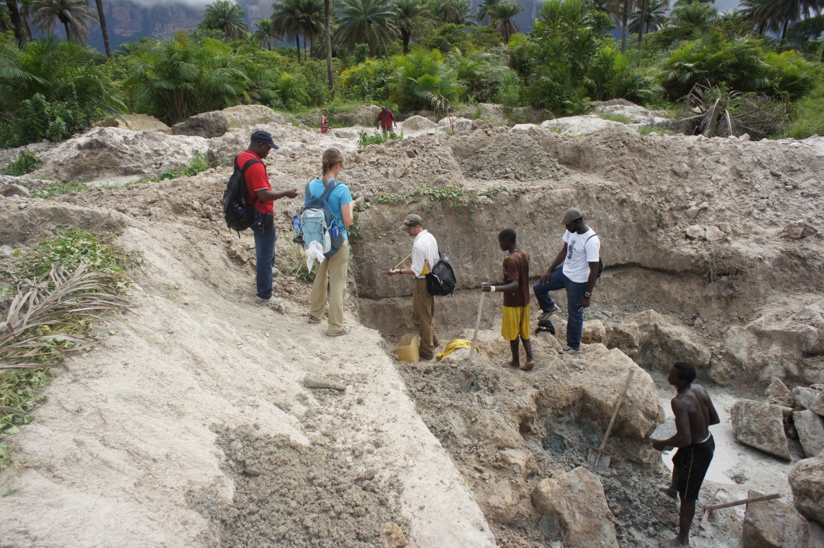 A small group of researchers and workers take measurements and samples from an open pit of dirt surrounded by jungle.