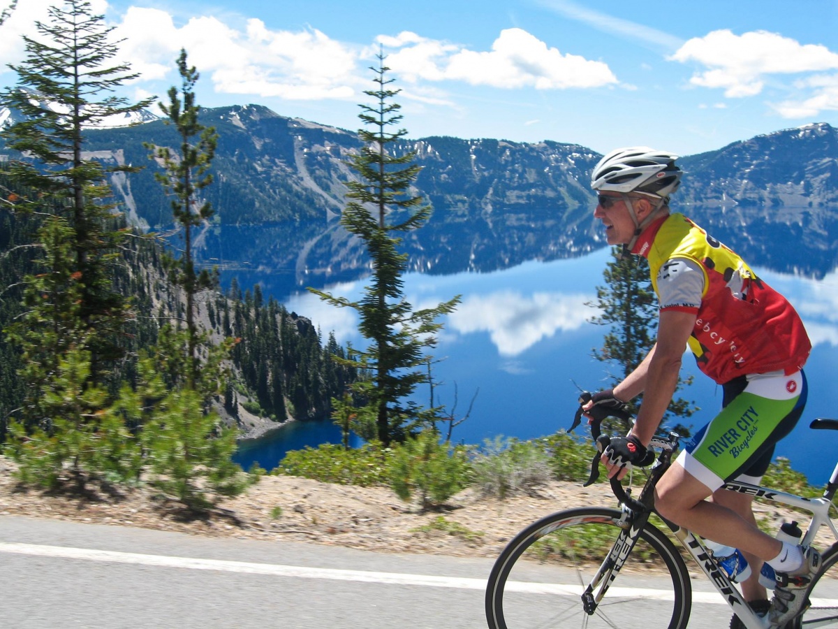 A thin, older white man wearing a helmet and bright biking clothes rides a bicycle on a road past a blue mountain lake.