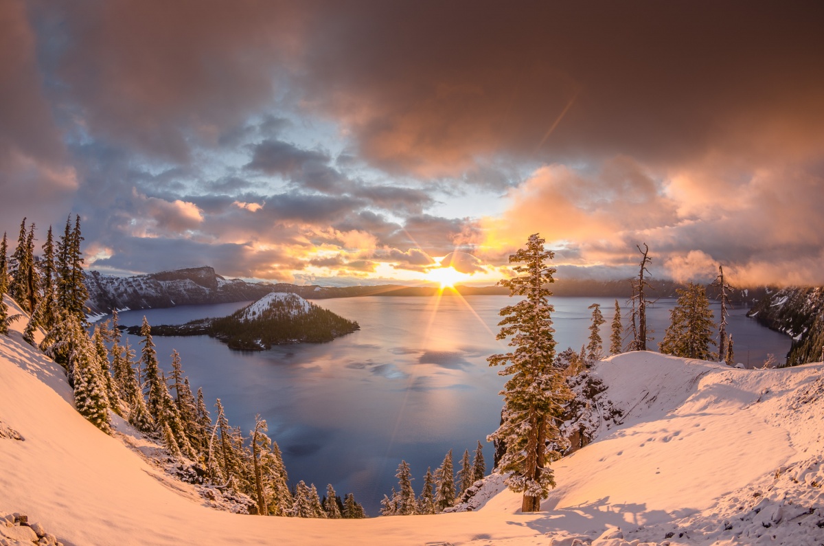 The sunrise peaks over the lip of a snow covered ridge that surrounds a circular mountain lake.