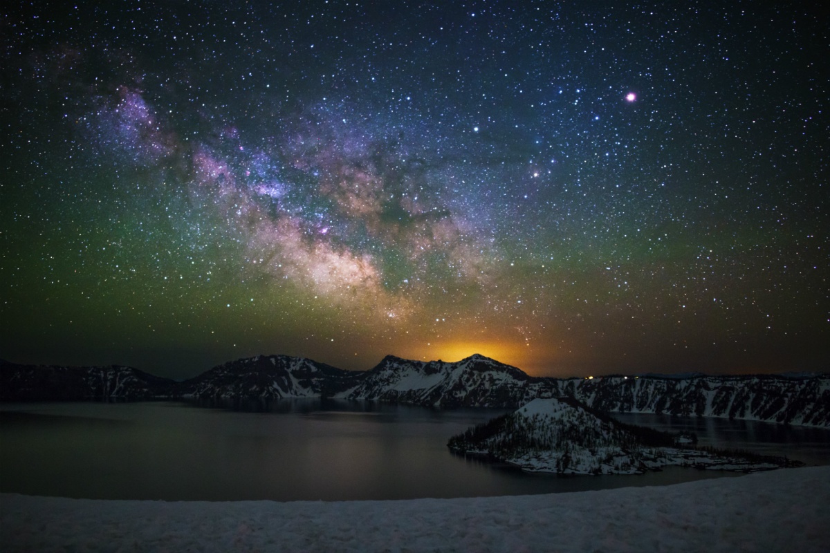 A dazzling night sky of stars and the milky way shines over snow covered cliffs sloping down to a wide circular lake. 