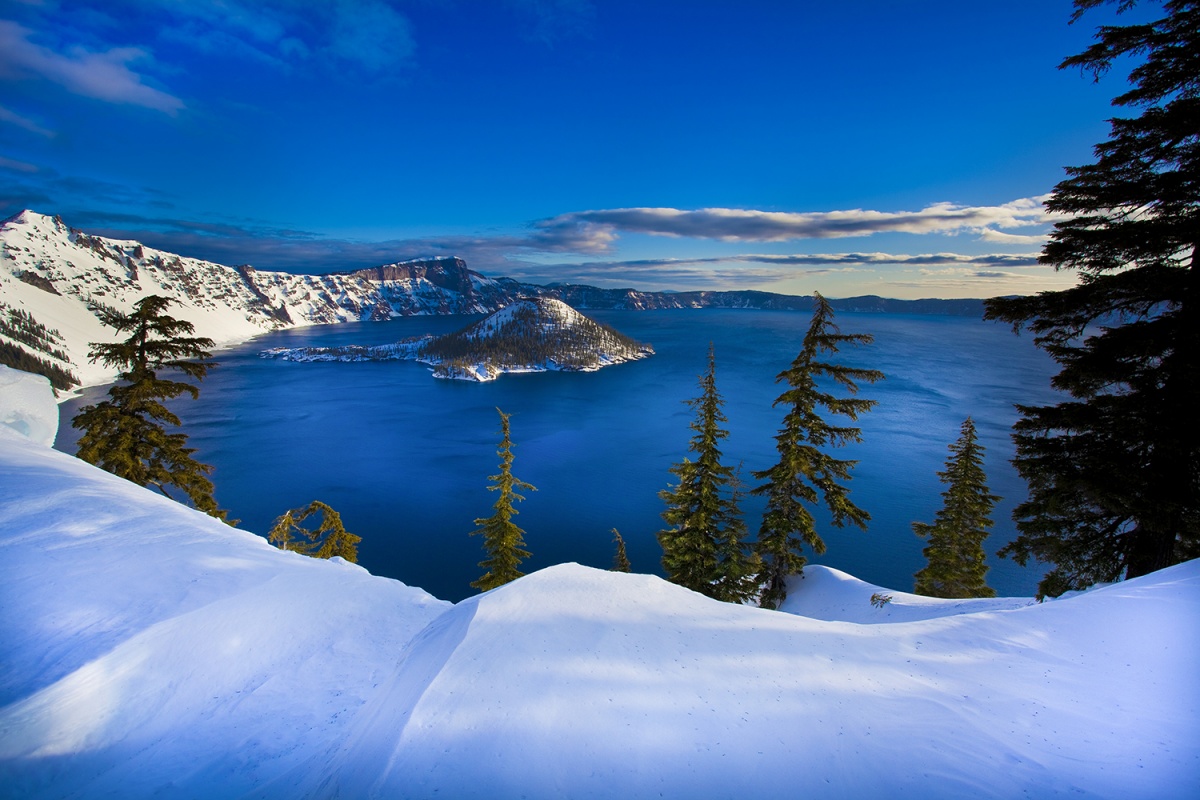 Crater Lake is a large, circular blue lake surrounded by a high rim of snow covered cliffs and a large pyramid shaped islands rises out of its calm waters.
