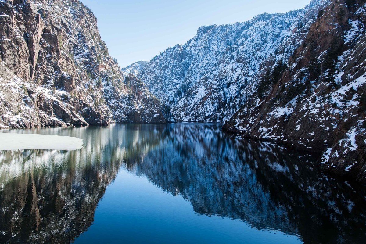 Snow covered walls of a canyon slope down to the flat, blue waters of a lake.