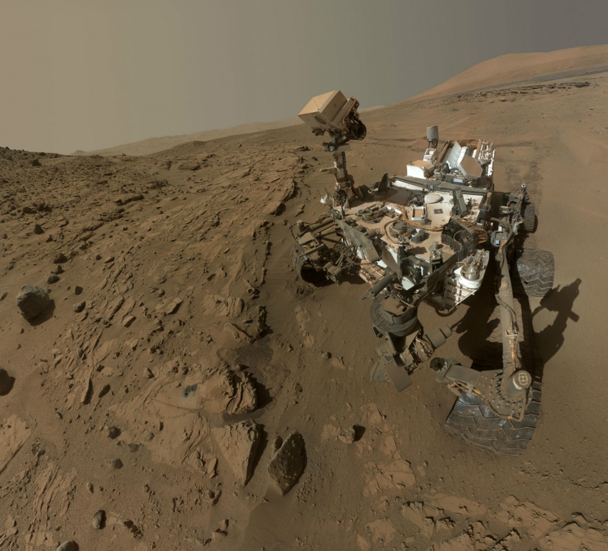 A small robot on wheels with metal arms and a camera mounted on a pole moves over dirt and rock on the surface of Mars.