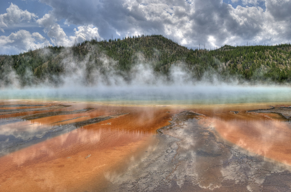 A colorful view of the prismatic spring with steam rising and mountains in the backdrop. 