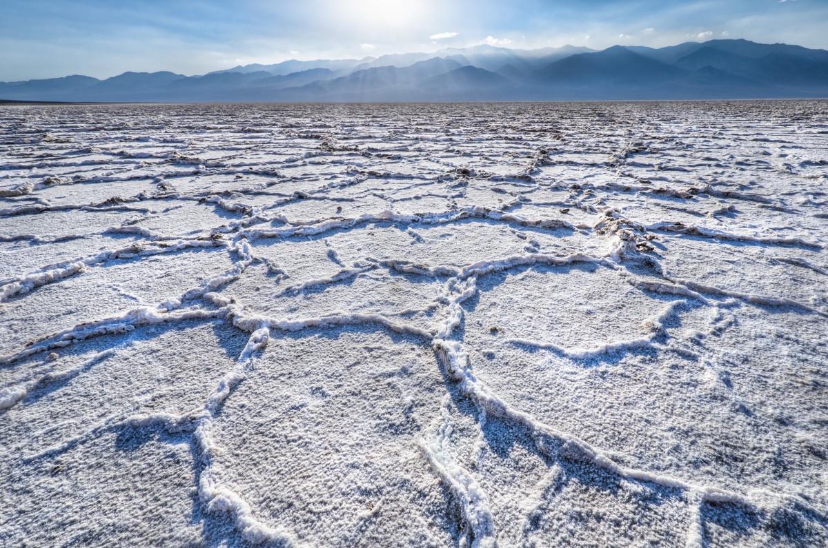 A flat desert plain showing ripples of white salt runs to a low range of mountains under a sunny blue sky.