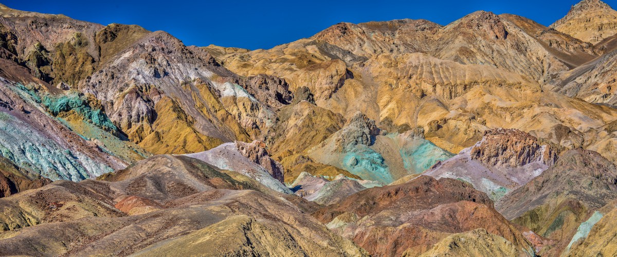 A wrinkled and ripple mountainside shows scattered deposits of blue, yellow and pink minerals.