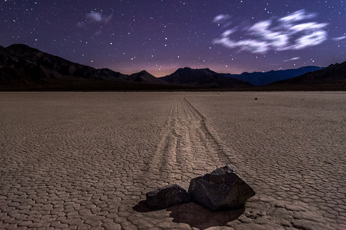 Two rocks sit at the end of long drag marks on a flat desert plain under a starry night sky. 