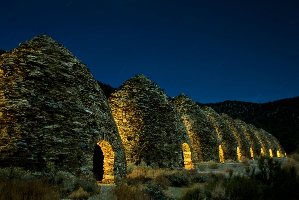 Nine rough stone cone shaped buildings with low arched doors stand in a line on a grassy plain under a night sky.