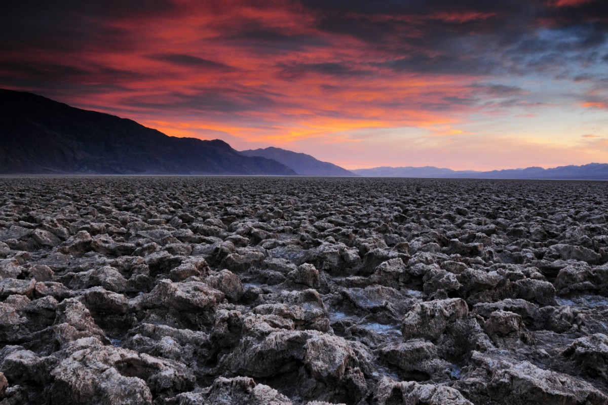 A flat desert plain bordered by mountains is covered in low jagged rocks under a pink sky.
