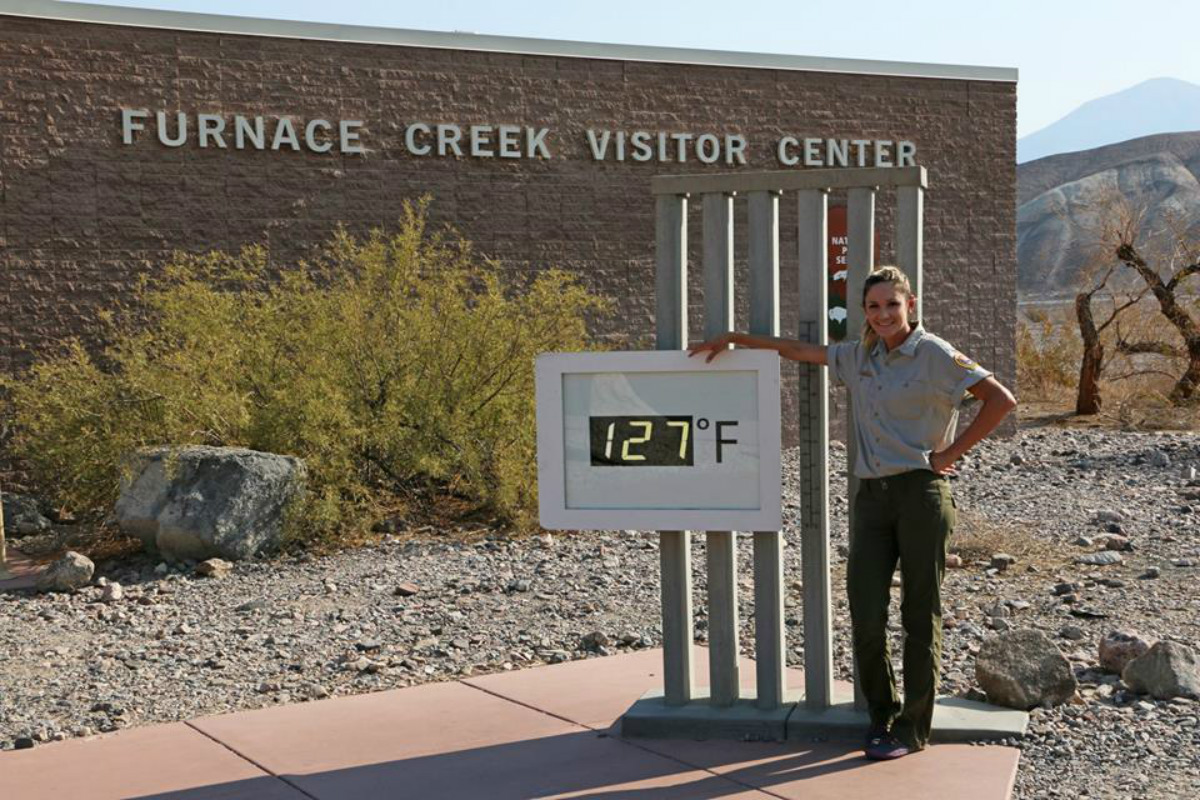 A young woman stands next to an outdoor digital thermometer that shows 127 degrees.