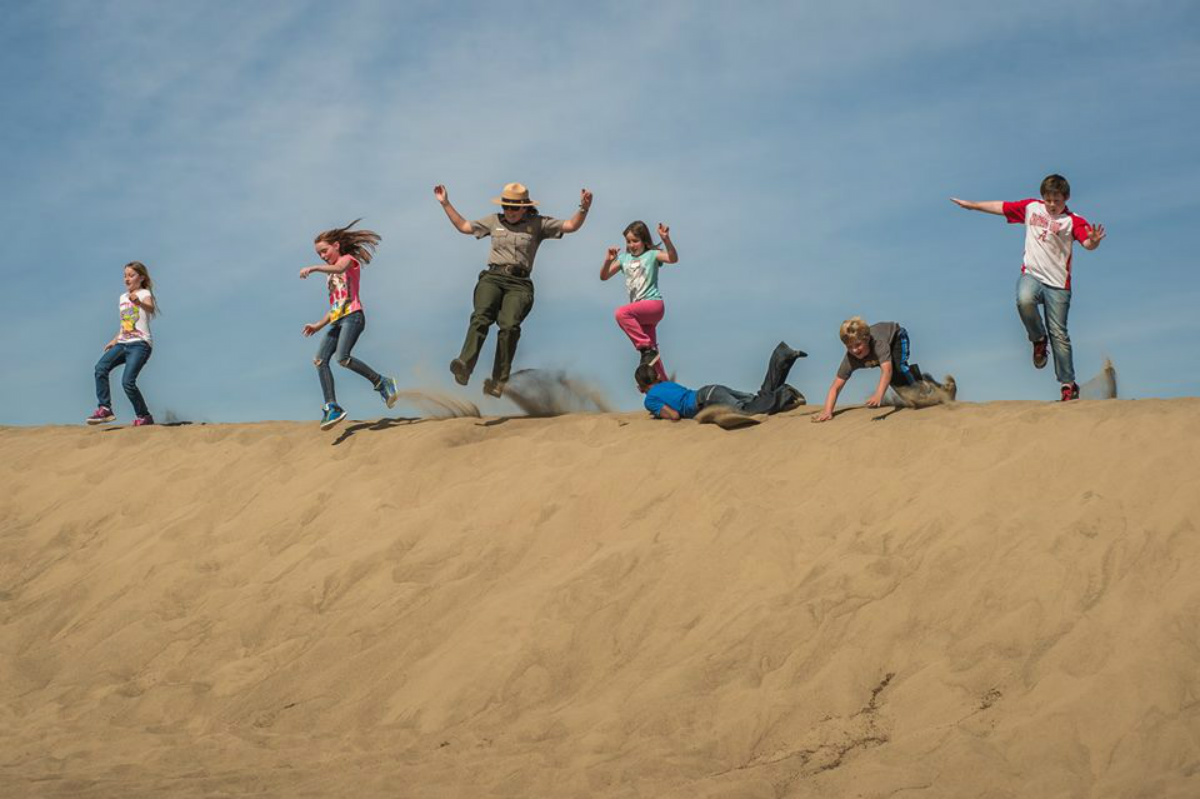 A female park ranger runs down a large sand dune with a group of kids.