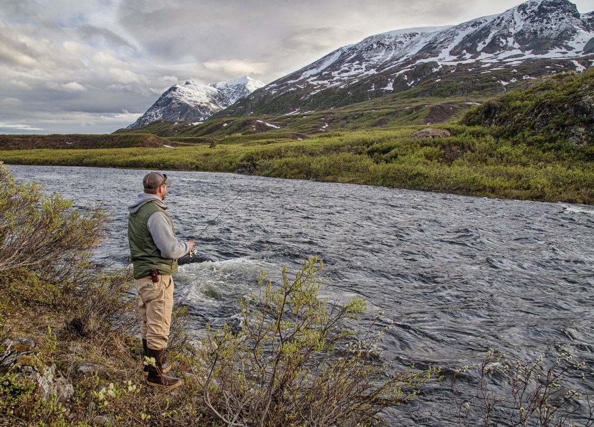 A man holds a fishing rod on a riverbank with a snow capped mountain in the background.