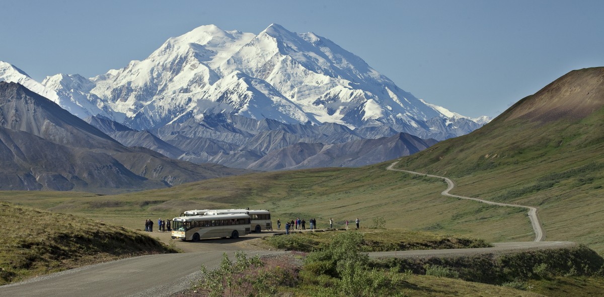 Two tour buses parked next to a road with people standing in the field next to them looking up at a huge snow covered mountain.