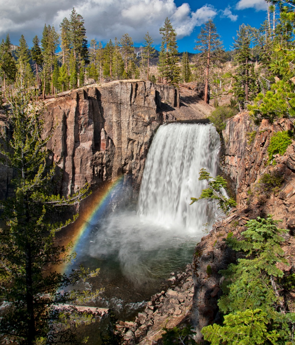 A waterfall flows from high rocky cliffs surrounded by bright green trees. A blue slightly cloudy sky shines over the scene, and a small rainbow frames the water.