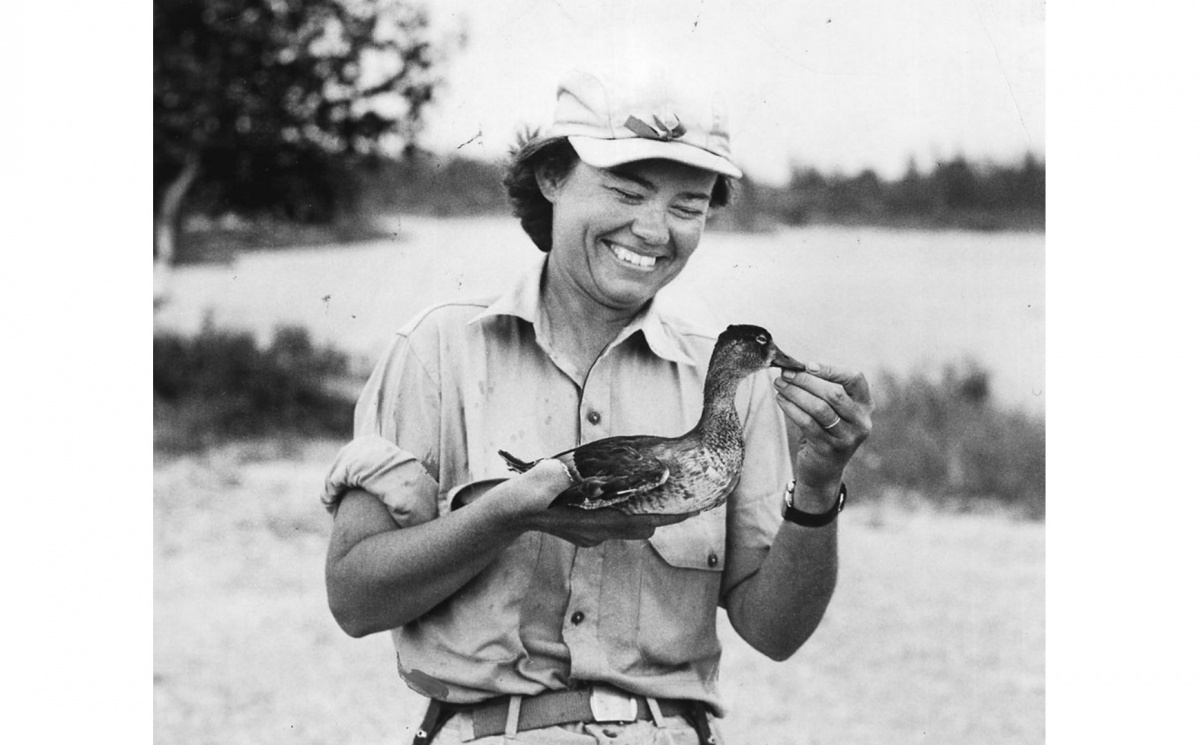 A black and white photo of a woman, Elizabeth Beard Losely, holding a bird in an open field and laughing.