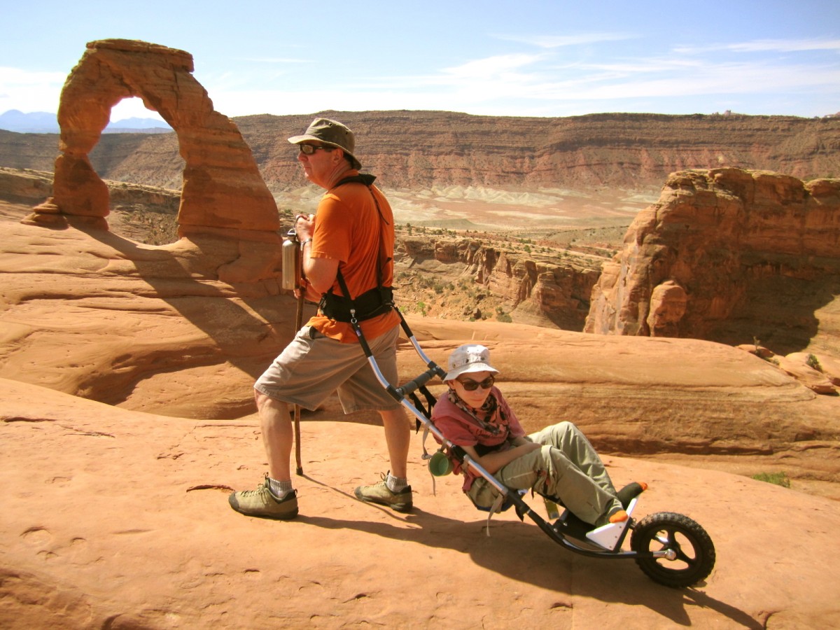 A young man with a disability sits in a modified wheelchair pulled up a stone plateau by an older man in outdoor gear.