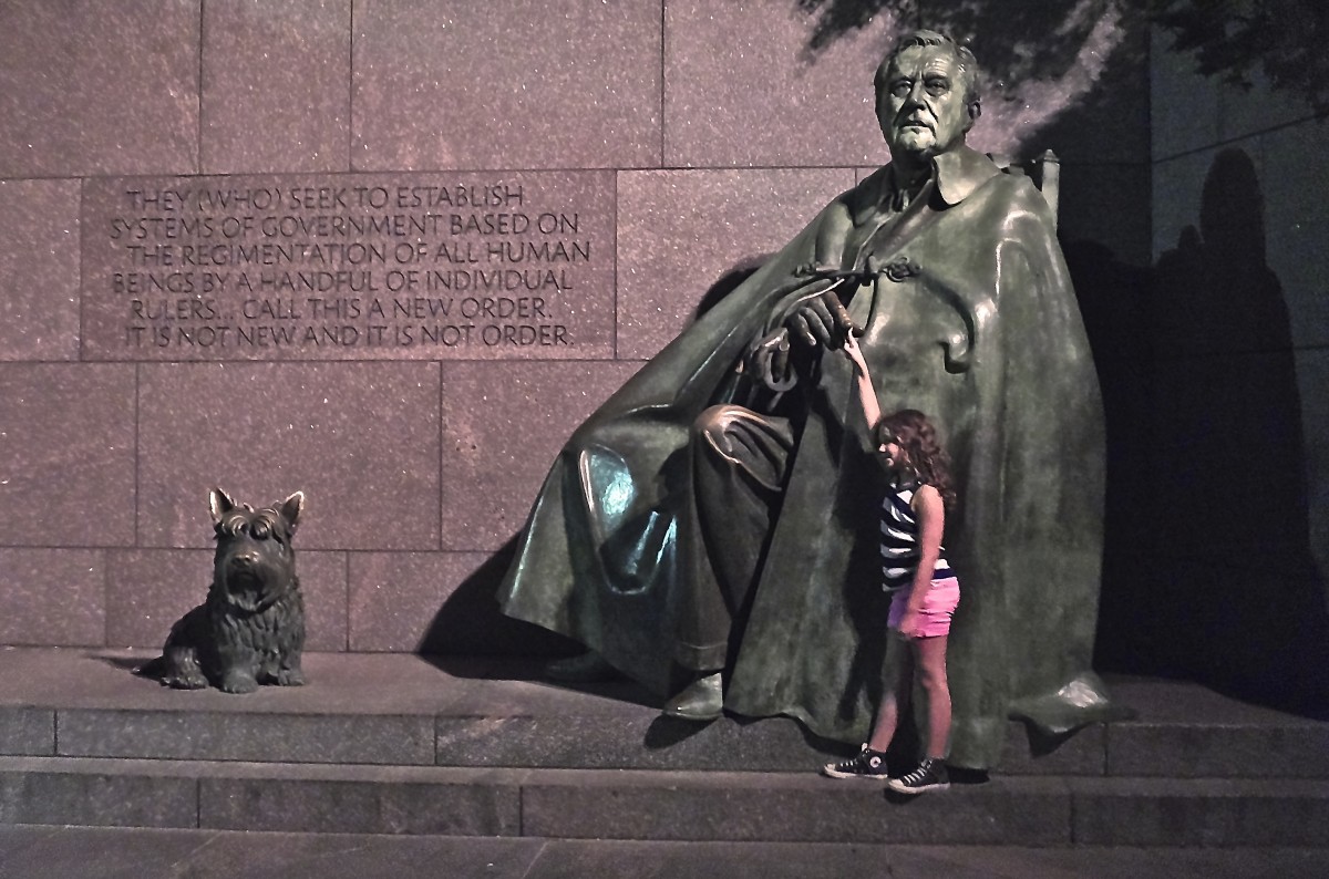 A young caucasian girl stands on a step and reaches up to touch the hand of a large green-bronze statue of FDR seated in a chair with his dog by his side.