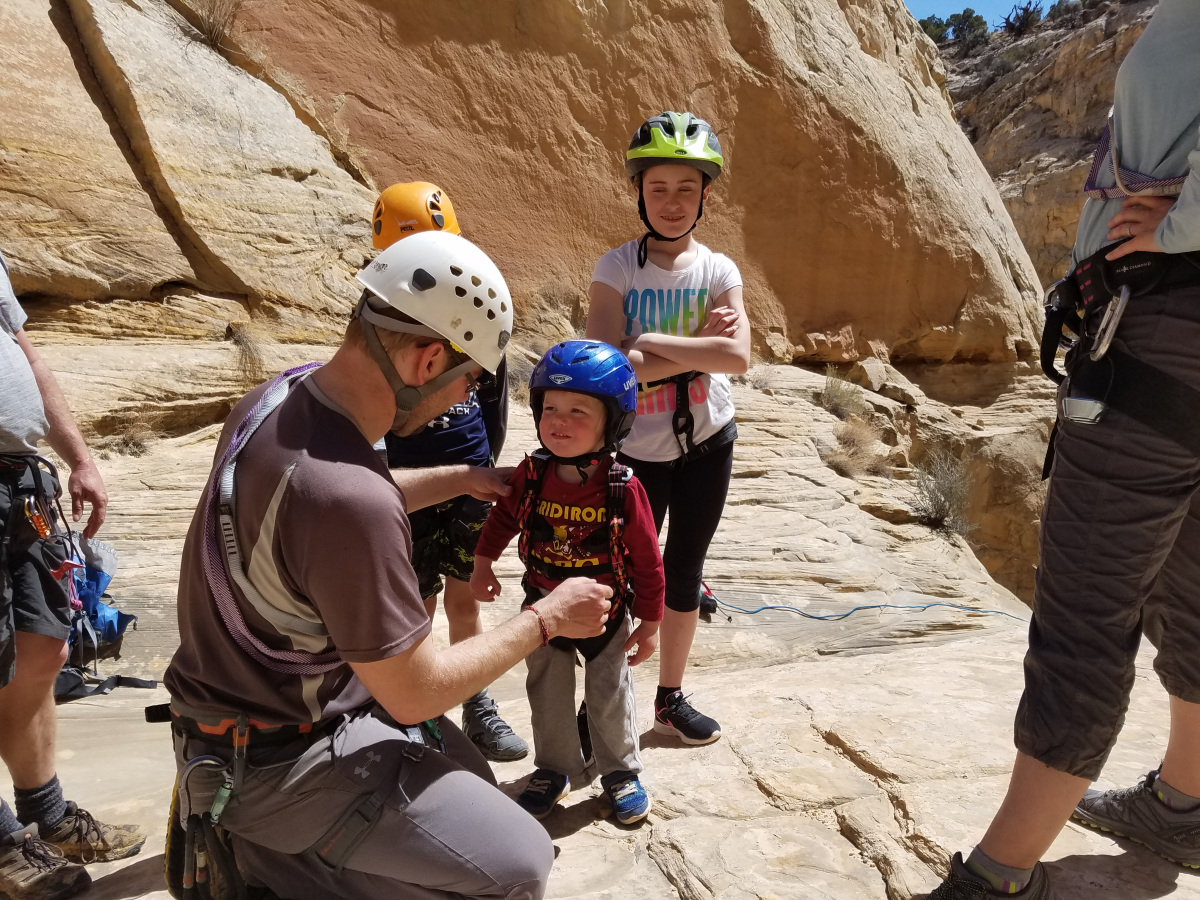 A man helps a little boy put on a climbing harness while other kids look on while standing next to a rock wall.