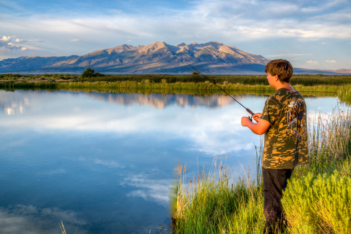 A young white man fishes in a still pond with a large mountain in the background.