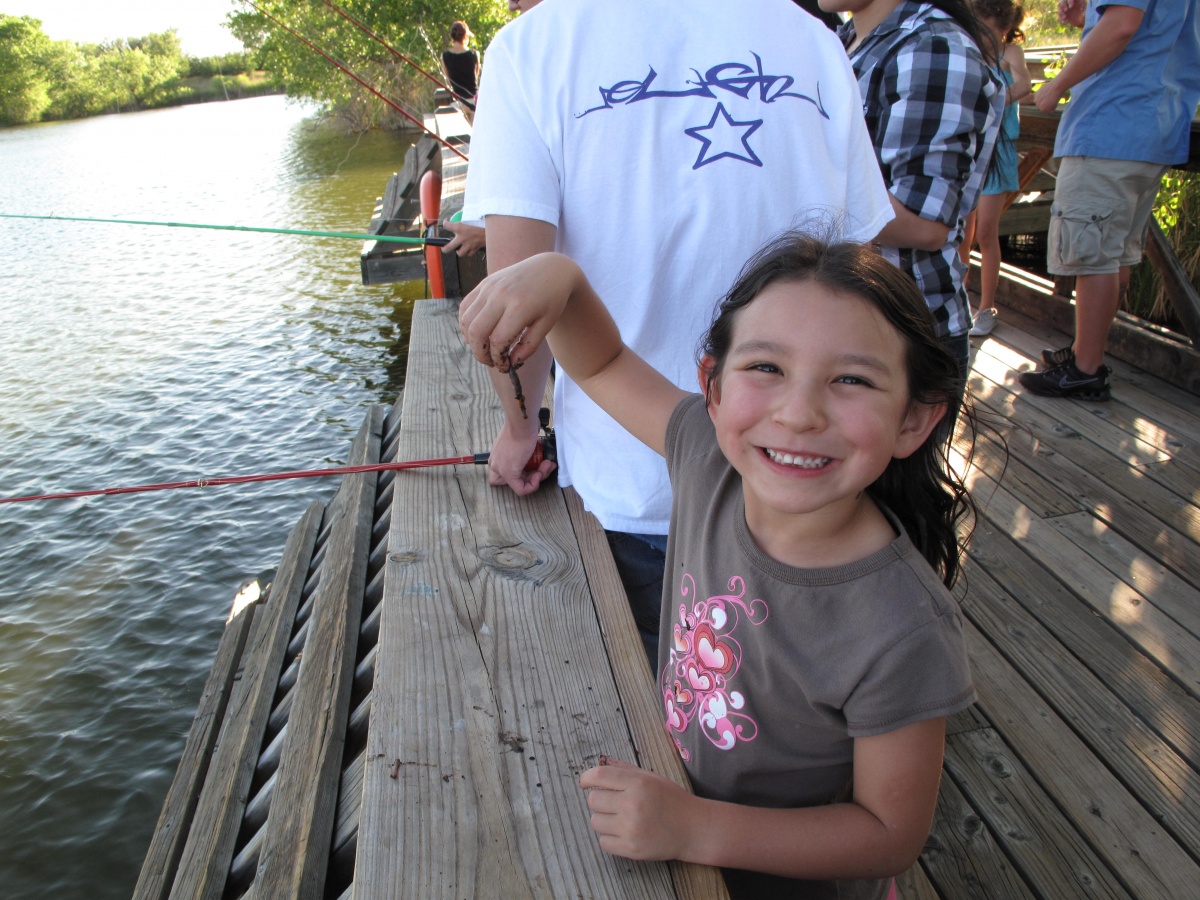 A little girl holds up a worm while standing on a dock with people fishing in a river.