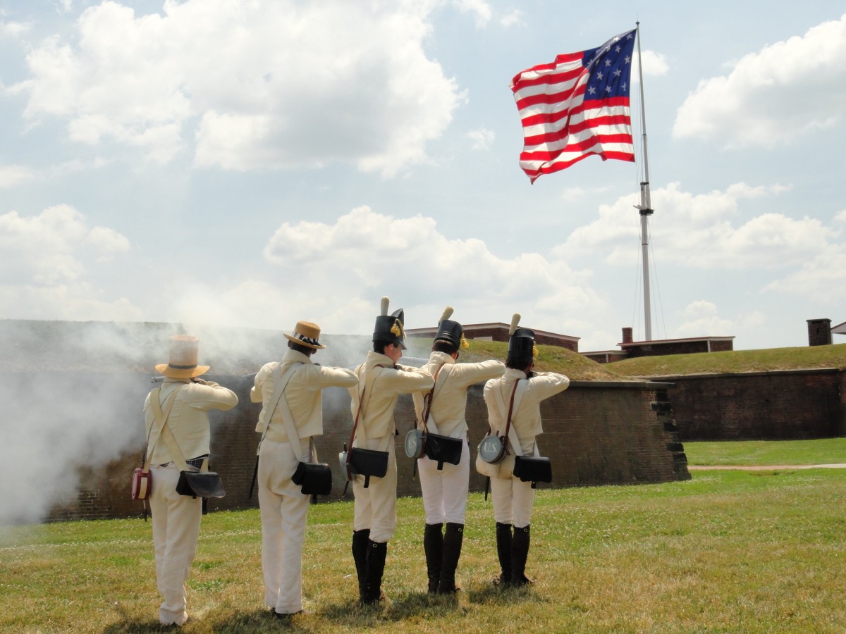 Five living historians stand tall and fire replica muskets toward a brick wall as smoke wafts from their weapons and the American flag waves above.