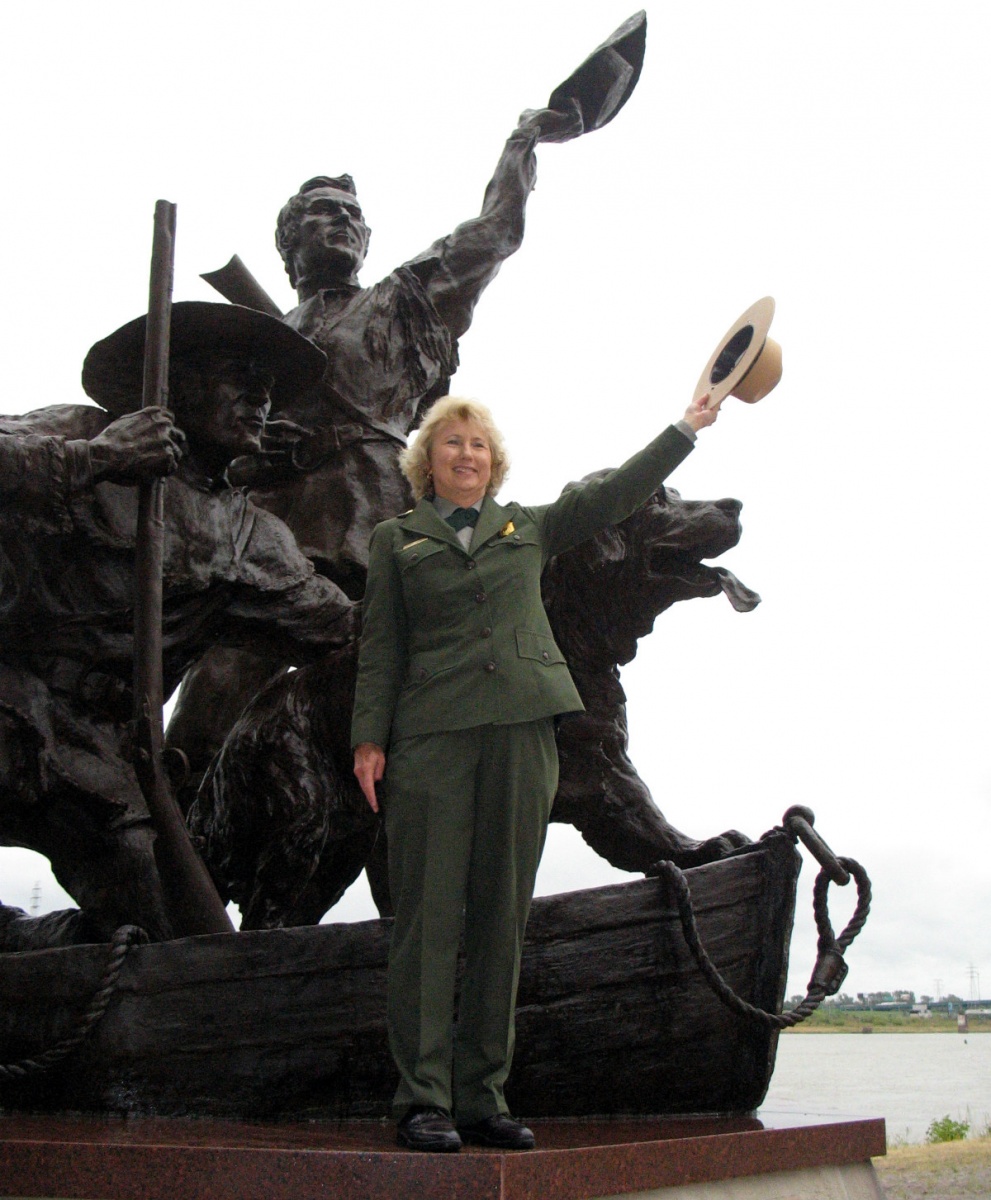 Fran Mainella -  a woman with blond hair wearing a national park service uniform -  stands in front of a statue of two men and a dog holding her hat in the air.