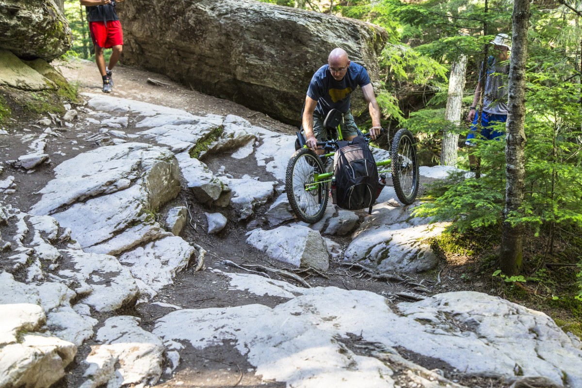 A bald caucasian man carefully maneuvers an offroad wheelchair over rocks on a trail through the forest.