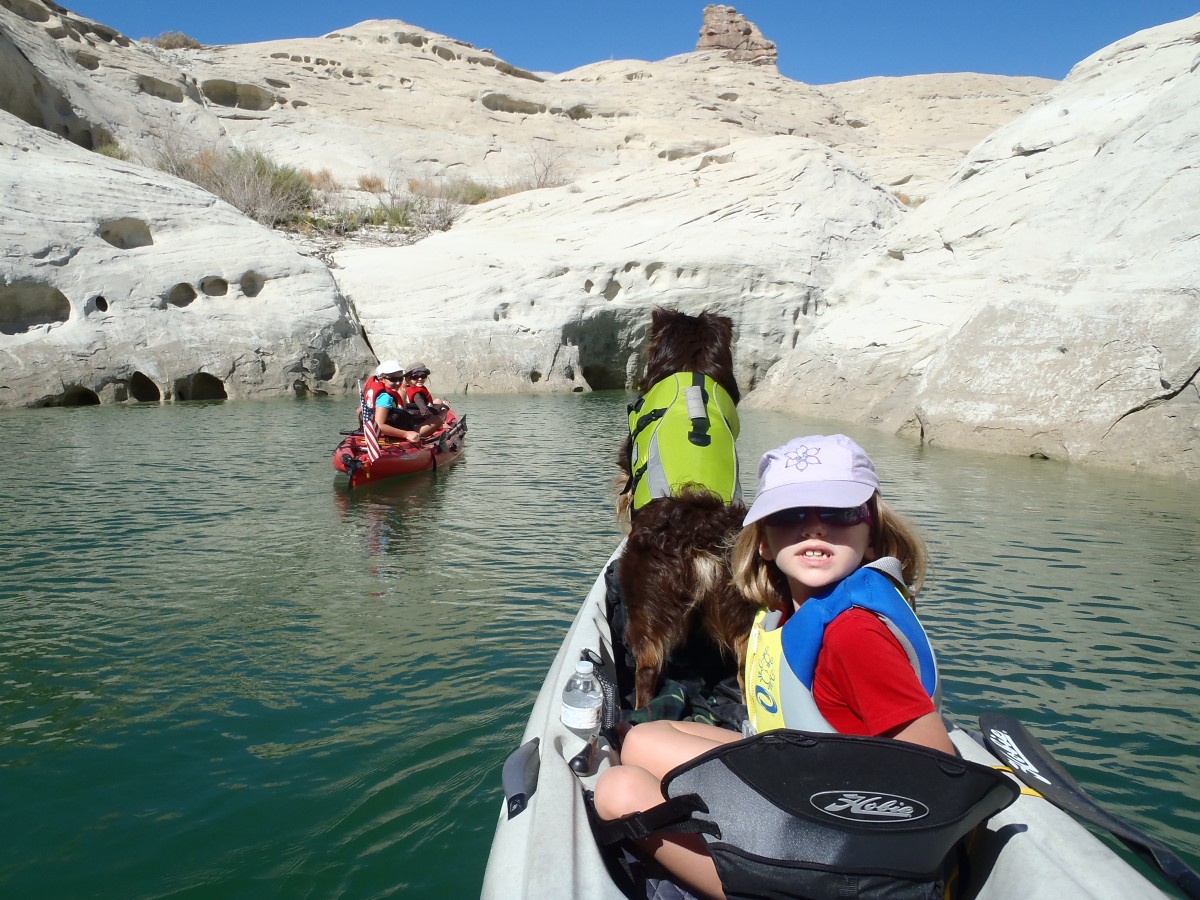 A little girl and a brown dog in a life preserve sit in the front of a kayak moving across the water towards another kayak floating next to a wall of white stone cliffs.