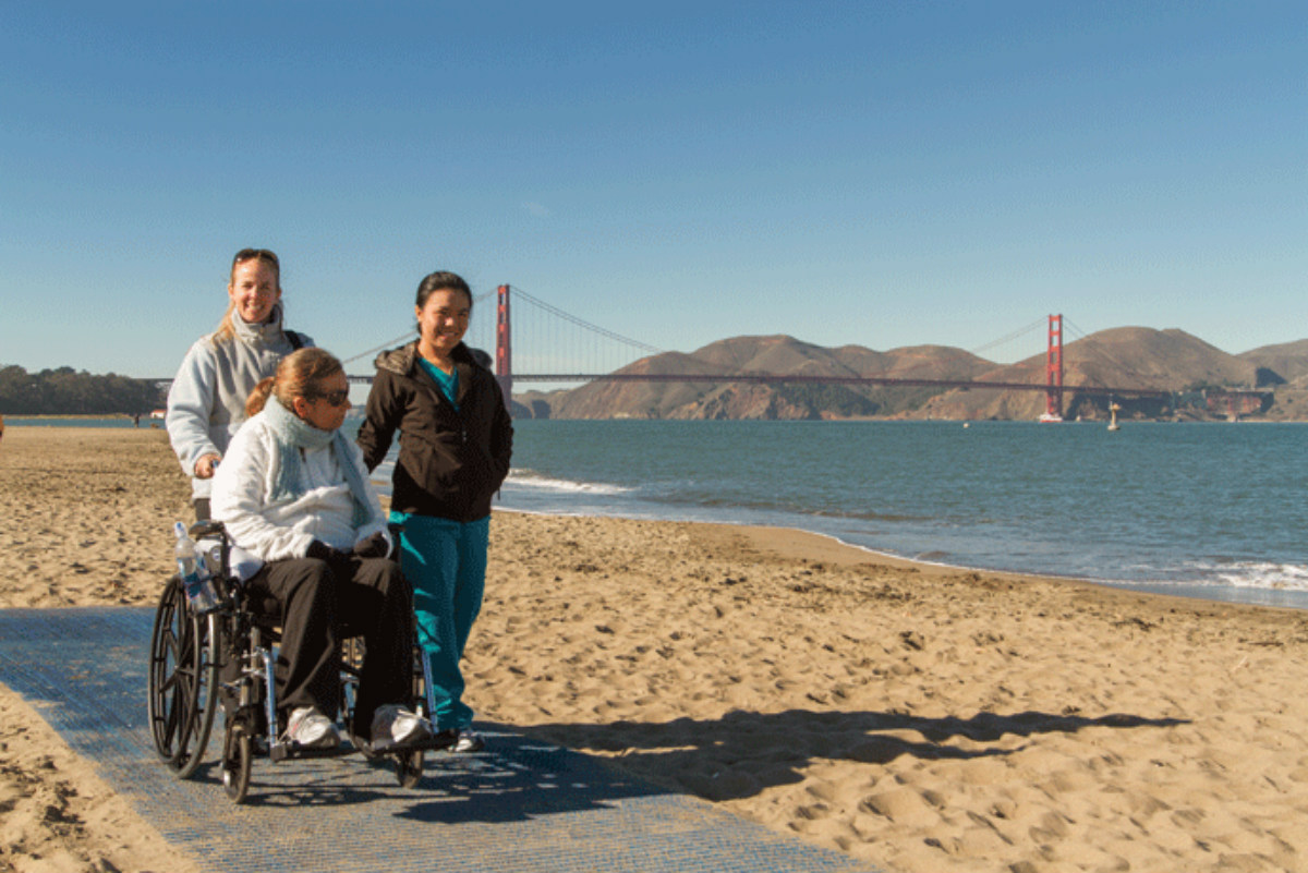An older blond woman in a wheelchair is pushed on a flat mat laid on the beach by two younger women with a view of the golden gate bridge in the background.