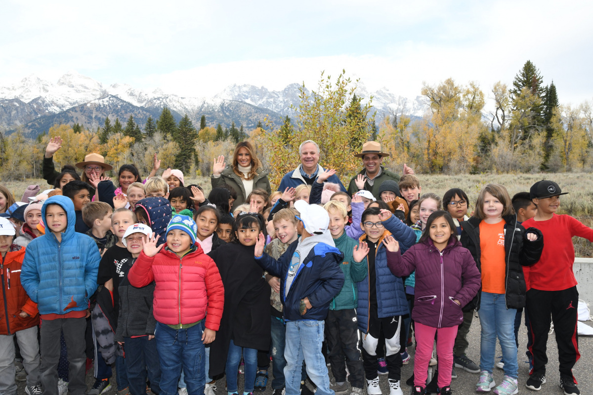 A large group of kids wave at the camera and stand with Secretary Bernhardt, First Lady Trump and some park rangers with a mountain range in the background.