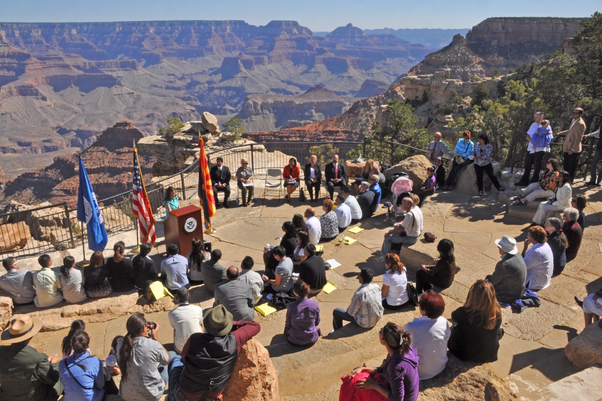 A large group of people listen as an official speaks at a ceremony on the rim of the Grand Canyon.