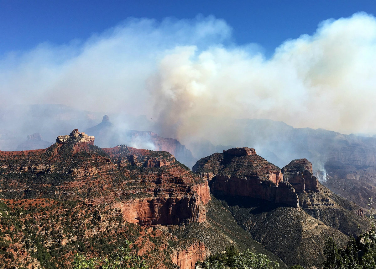 White-yellow smoke billows from behind layers of orange canyon wall covered in brush
