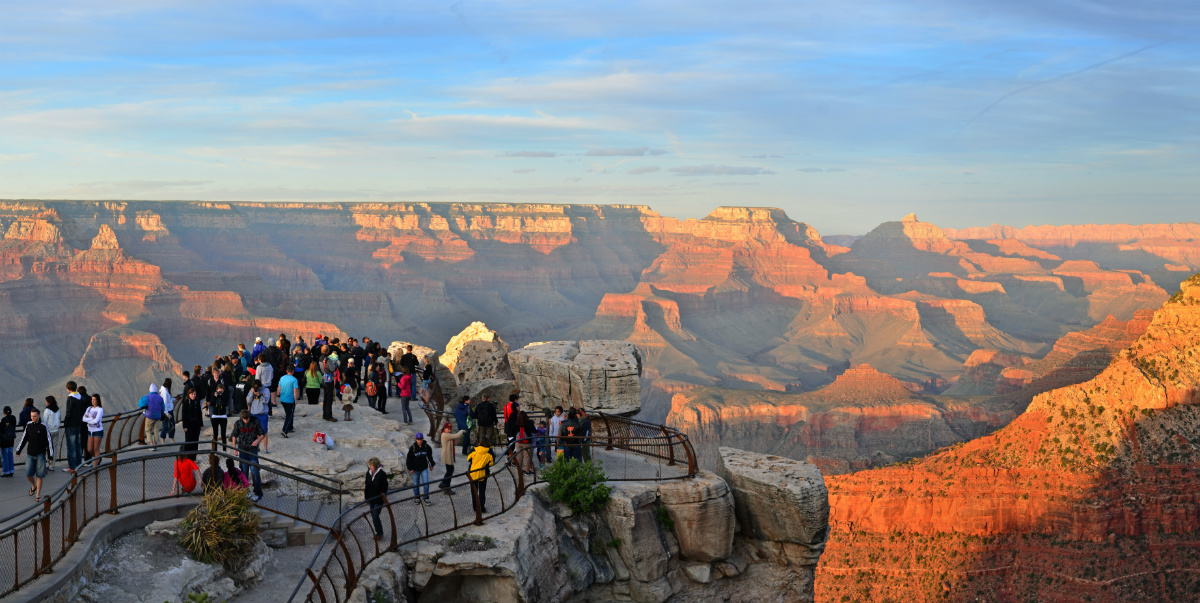 A crowded fenced viewpoint overlooks the Grand Canyon as the sun sets, hitting the tops of the Canyon walls