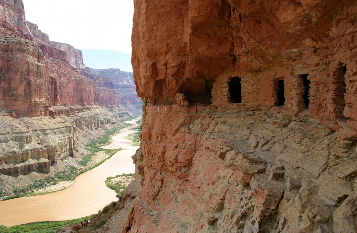 On the right side of the picture is a rough wall of deep red with caves lined up, carved into the Canyon wall. On the left is another wall of a lighter orange shade towering over a muddy Colorado river 