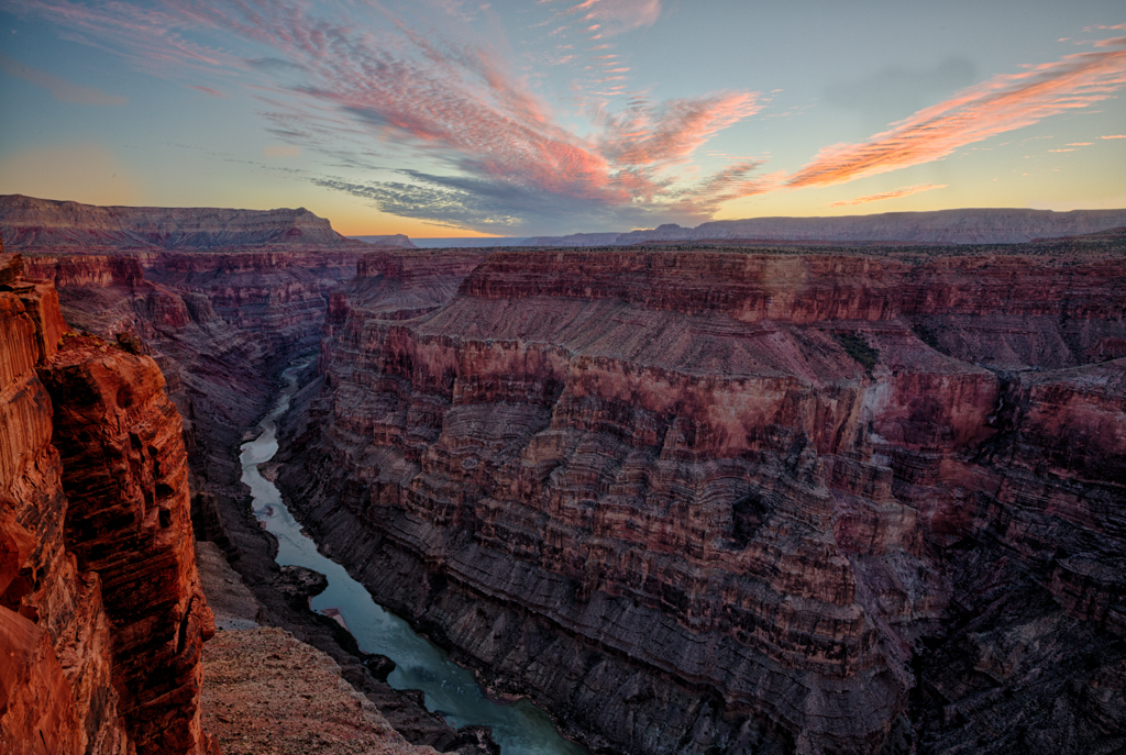 A fiery sun sets over the canyon with striking pink and purple clouds and layers of purple and red rock line the turquoise river below