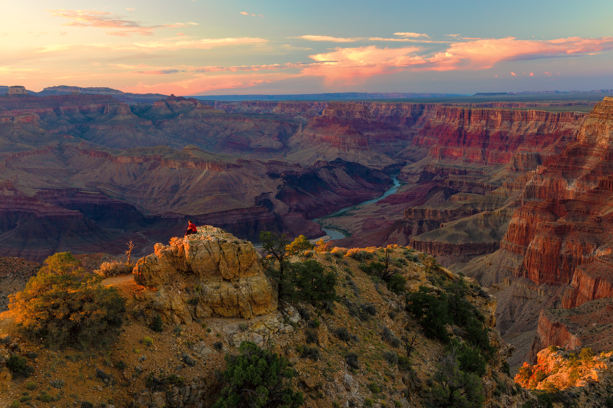 An off-screen sunset paints an elevated rock yellow as a man perched on top, looks into the distance. The canyon is a range of purple and oranges extending into the horizon and anchored by a brilliant blue Colorado river
