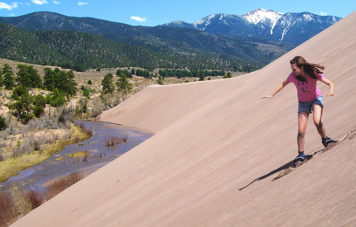 A girl sand boards down the side of a dune. A mountain and river are in the summer background.