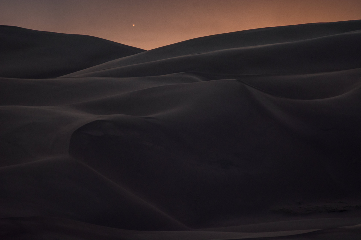 two large dunes overlapping at dawn. Mars is glowing in the sky with other stars.