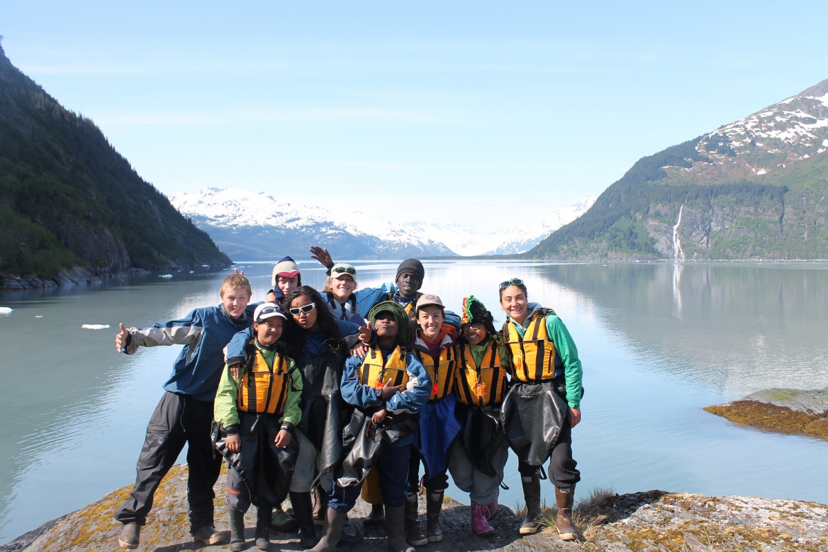 A group of young people in kayaking gear pose in front of a calm lake surrounded by mountains.