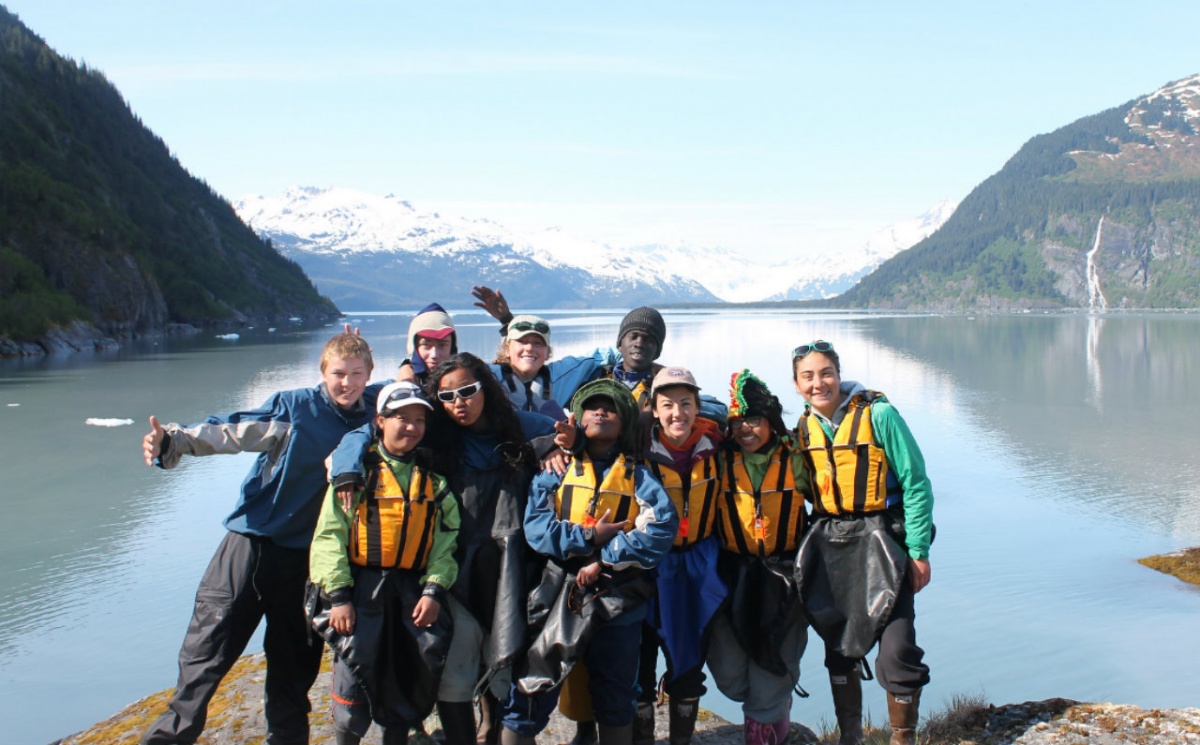 A group of young people dressed in kayaking clothes pose together in front of a lake with mountains in the background.