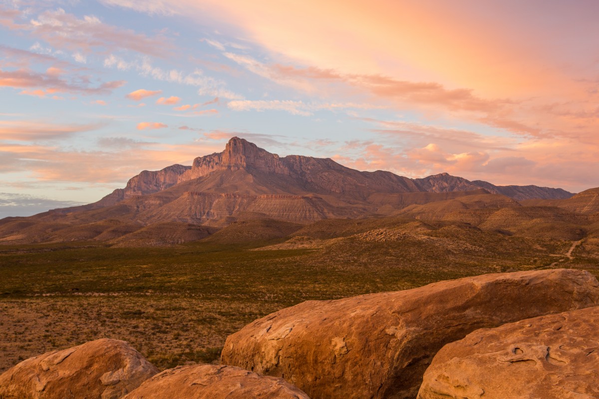 Rocky, tan mountains rise over sparse desert shrubs amidst a picturesque sunset with brilliant oranges and pinks.