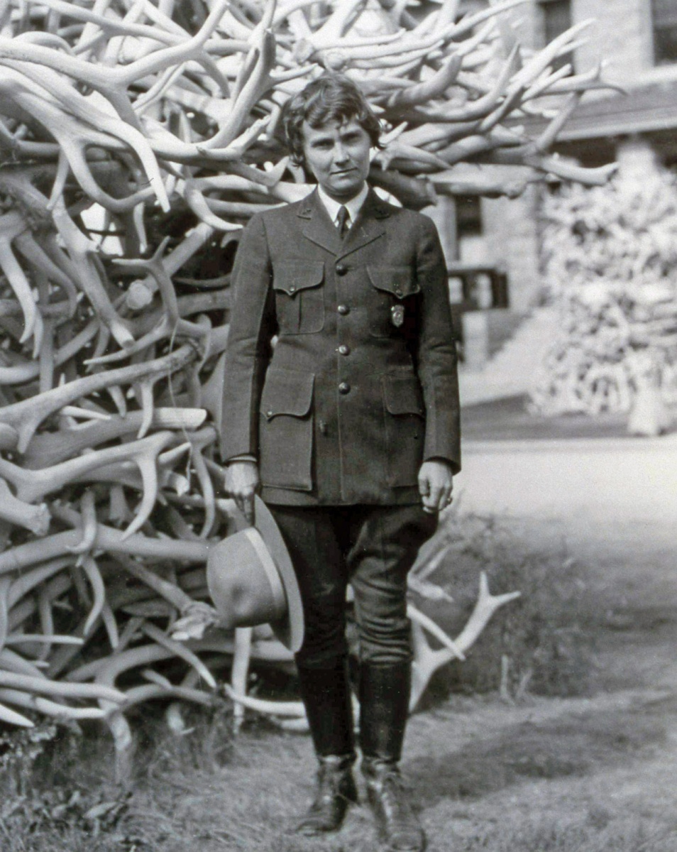 A historic black and white photo of Herma Baggley wearing her National Park Service uniform and standing in front of a tall pile of elk antlers.
