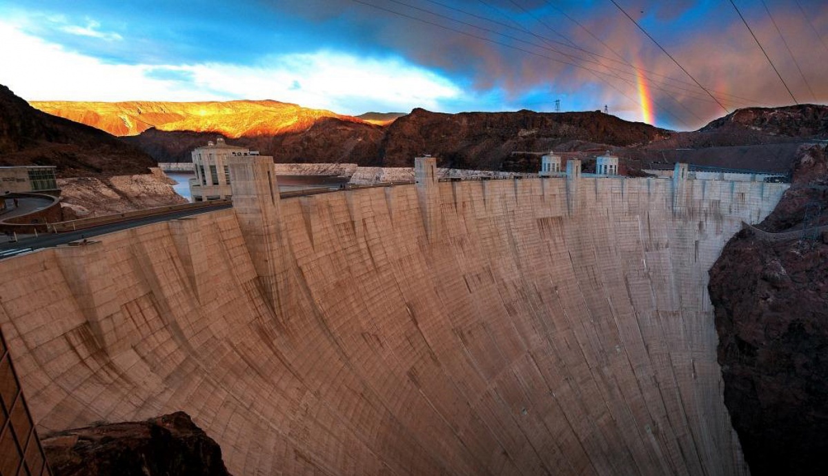 A rainbow hovers over a lake that fills a canyon behind a large concrete dam.