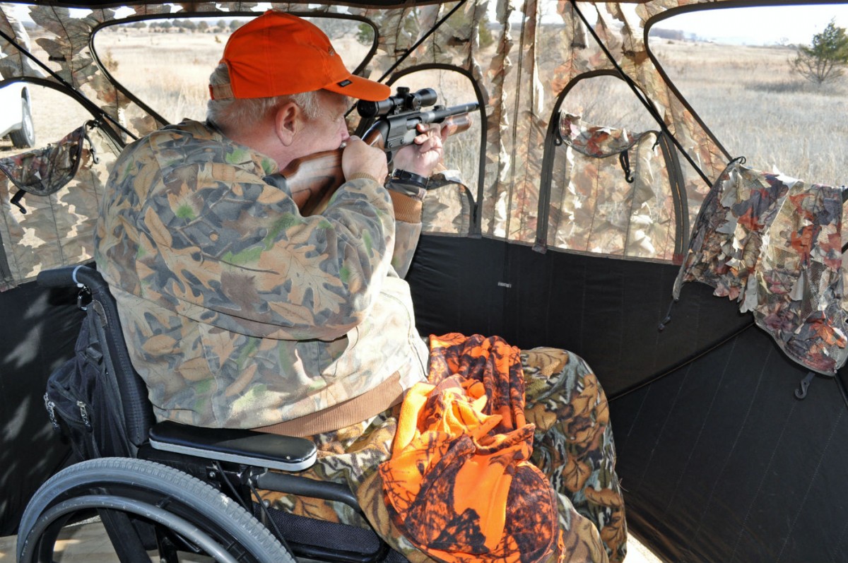 An older caucasian man in a wheelchair wears camouflage hunting gear and points his rifle out of a hunting blind toward a grassy field.