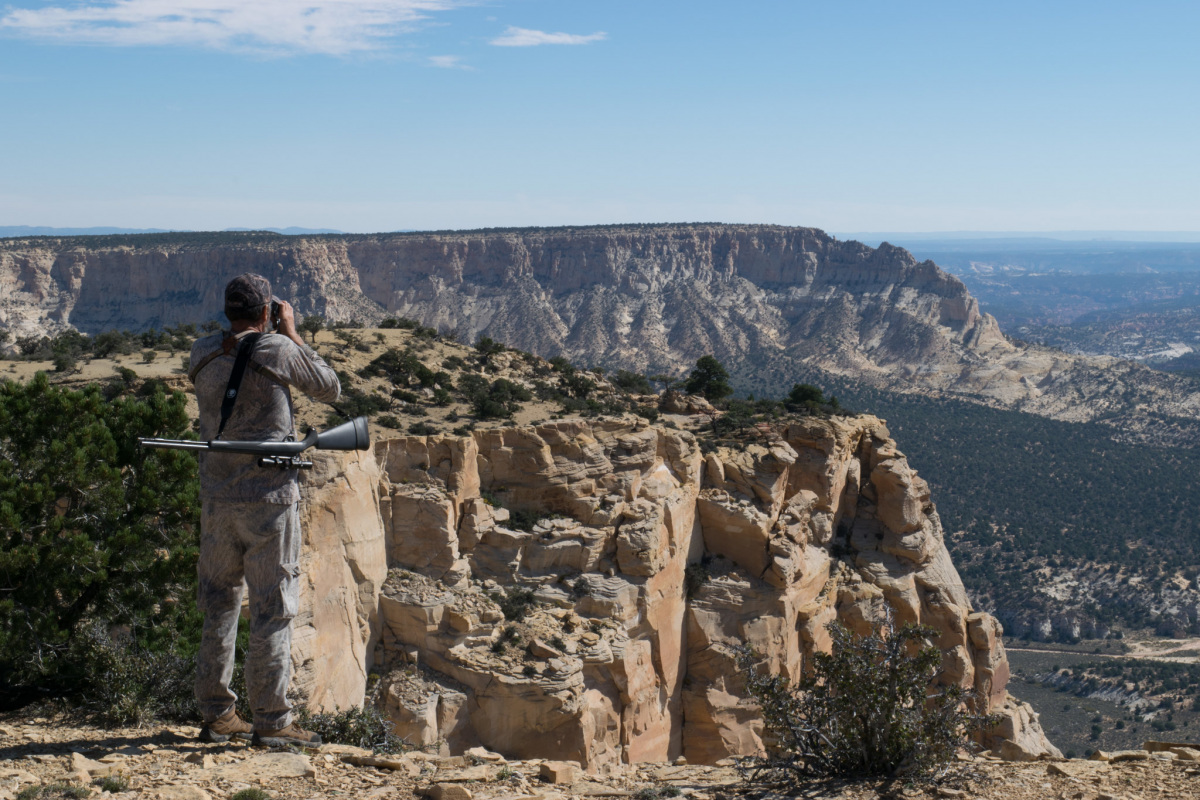 A man with a rifle slung over his shoulder stands on a cliff looking down into a valley with binoculars.