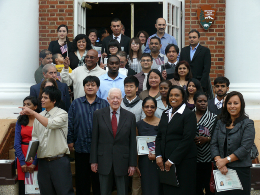 Former President Jimmy Carter stands with a group of new citizens on the steps of a brick building.