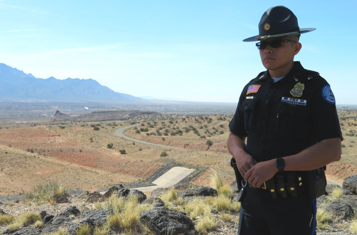 A Native American male police officer wearing his wide hat, uniform and sunglasses stands on a hill overlooking a grassy plain.