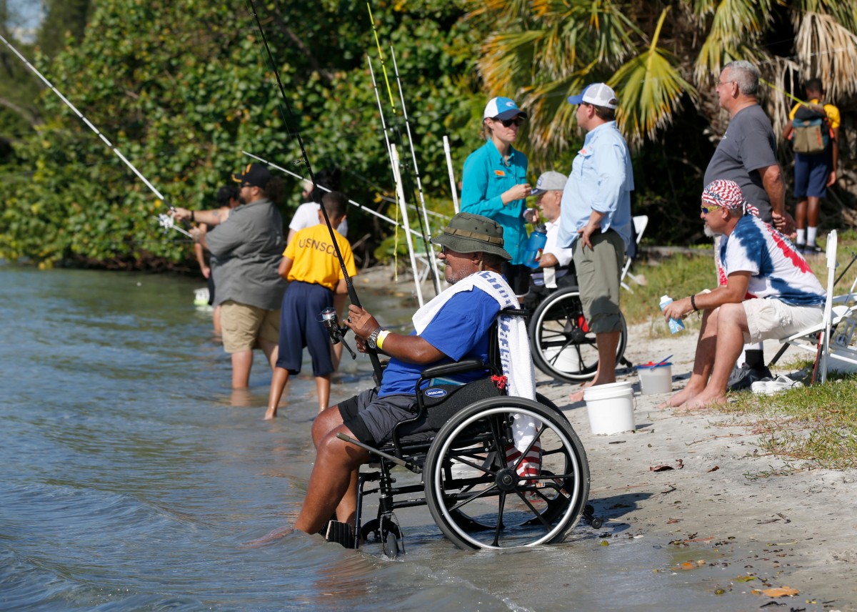 Several veterans relax on the sand at the water's edge, casting their lines into the clear water and socializing among family and friends with green bushes rising in the background.