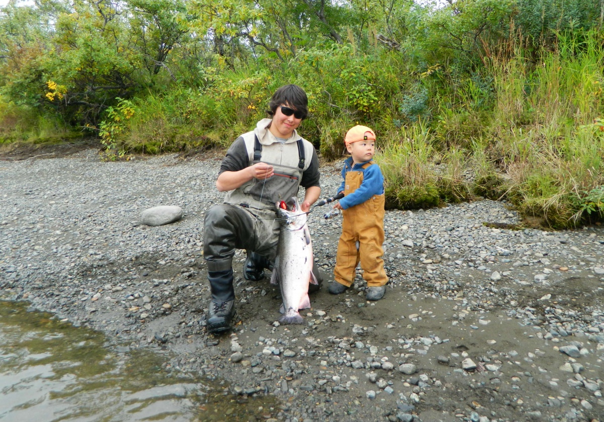 A young man and a boy hold a fish on the bank of a river in Alaska.