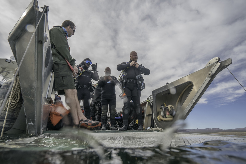 Three veterans stand next to the water on the edge of a boat's gray deck, adjusting their diving gear and goggles with blue skies and clouds high above.